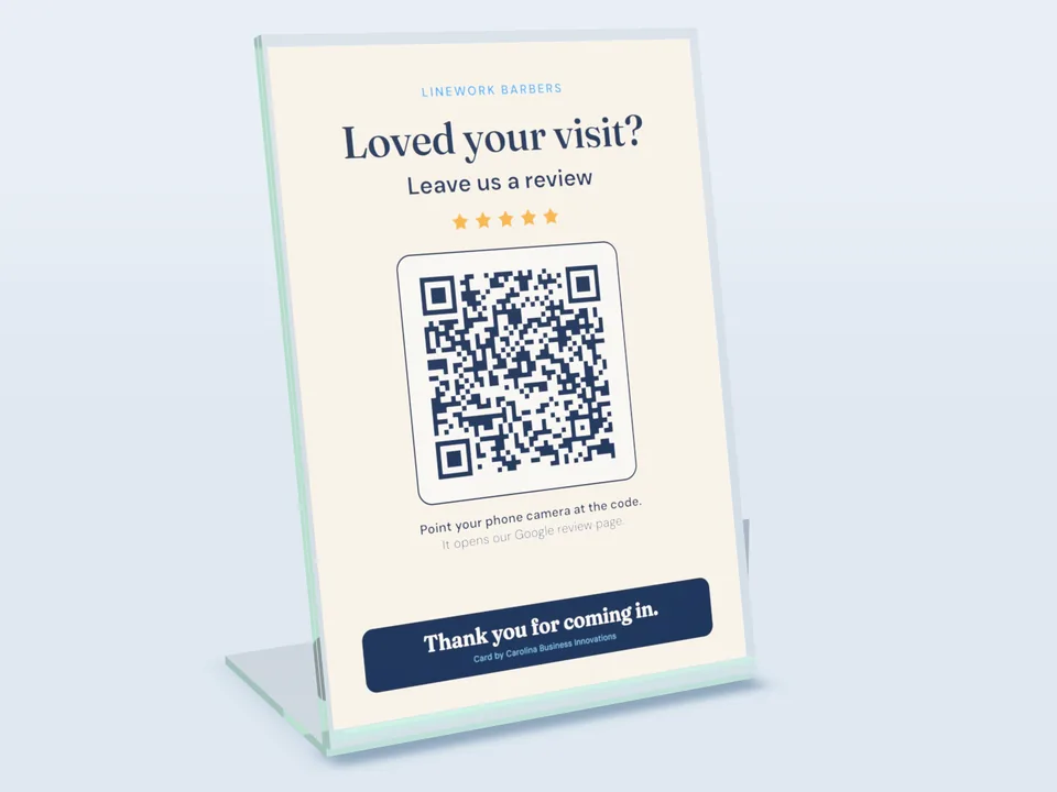 The Review Booster: a 5 by 7 inch counter card in a clear acrylic stand. It reads “Loved your visit? Leave us a review” above a QR code. Shown for a made-up shop.