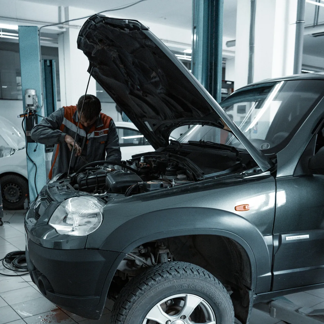 A mechanic works under the open hood of a grey SUV in a bright, clean service bay.