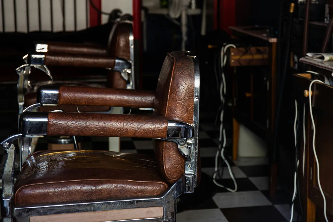 Two empty vintage barber chairs in brown leather and chrome, on a black and white checkered floor.