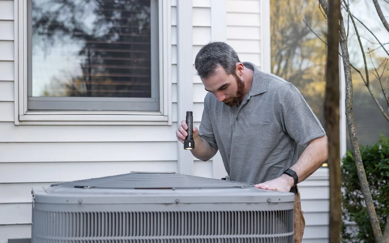 An HVAC technician works on an outdoor air conditioner unit beside a house.